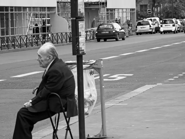The old man wearing suit is sitting in a chair and there are cars on the road behind him.