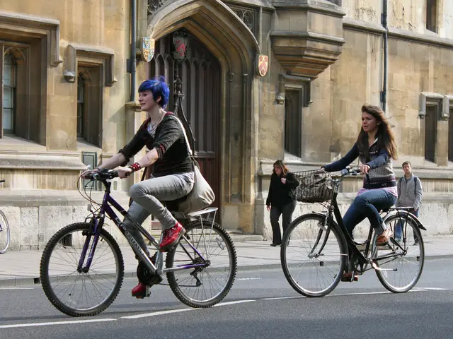 Two woman are riding a bicycles on the road and two people are walking on the footpath on the other...